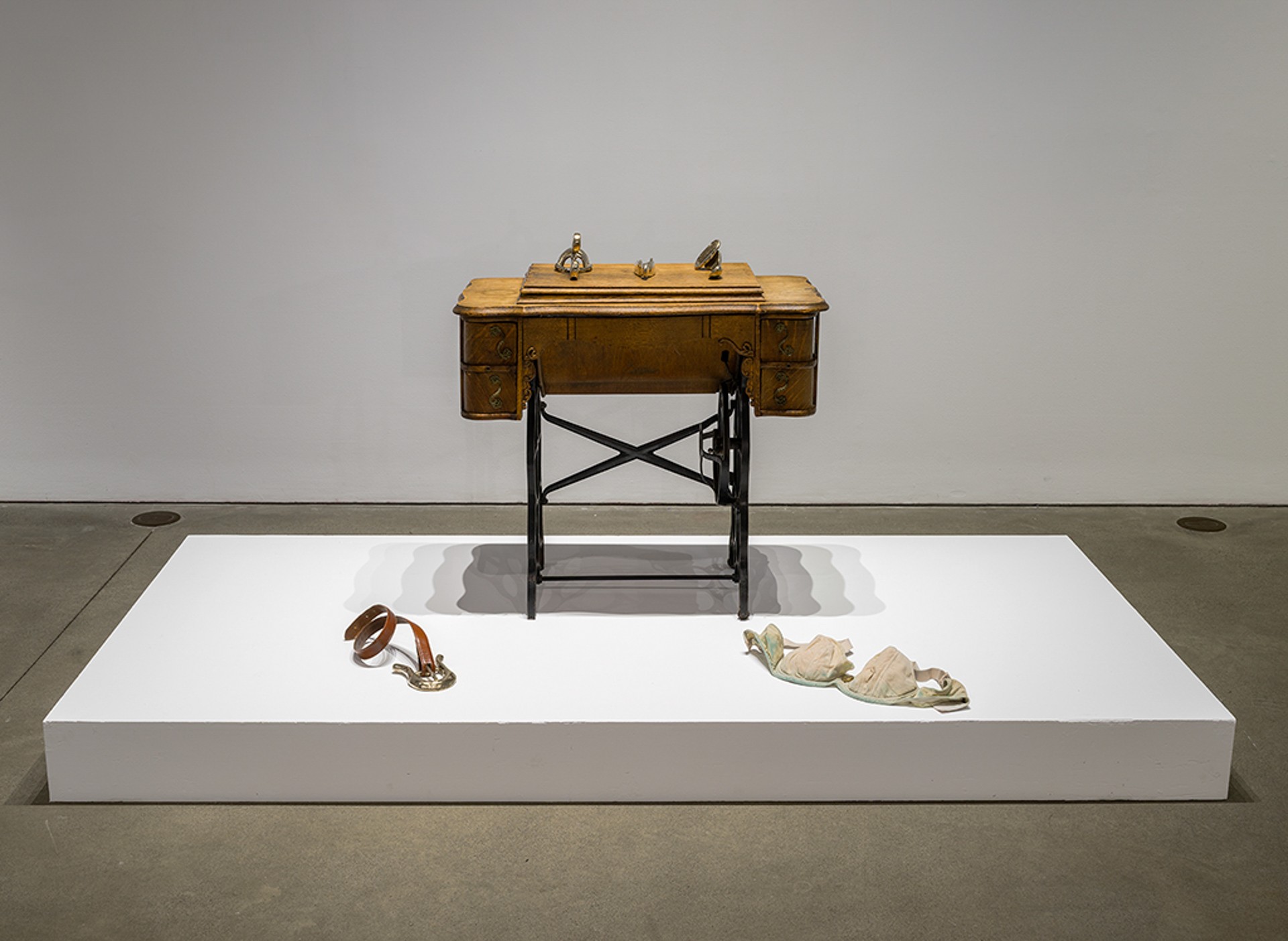 Ornate sewing table with three bronze objects laid on its surface and on the floor: a stained bra and a belt with an oversize buckle