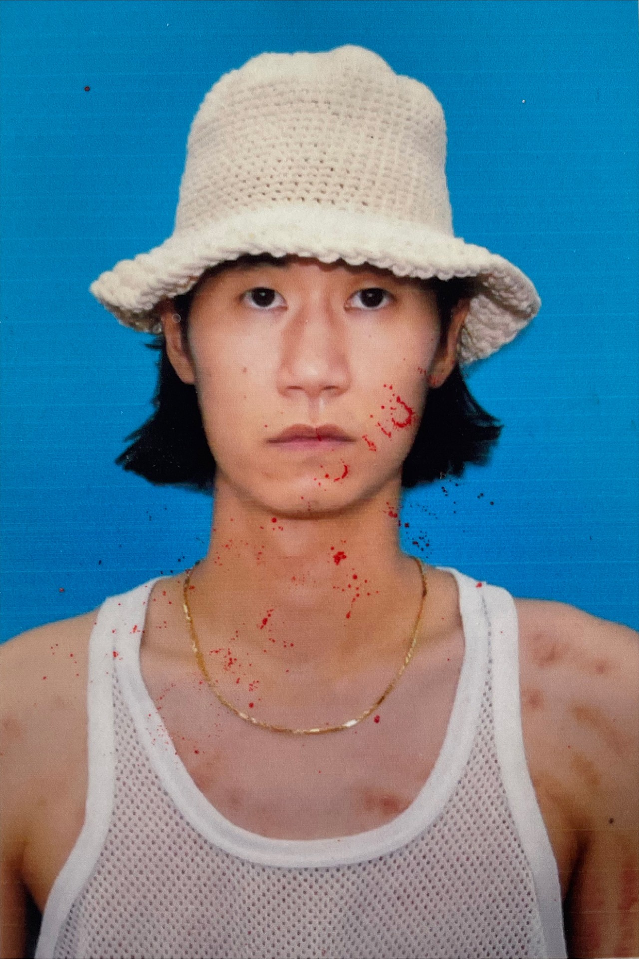 Portrait de l'artiste vêtu d'un haut de mailles blanc, d'une fine chaîne en or et d'un chapeau blanc. Une éclaboussure de sang est ajoutée numériquement sur son visage.