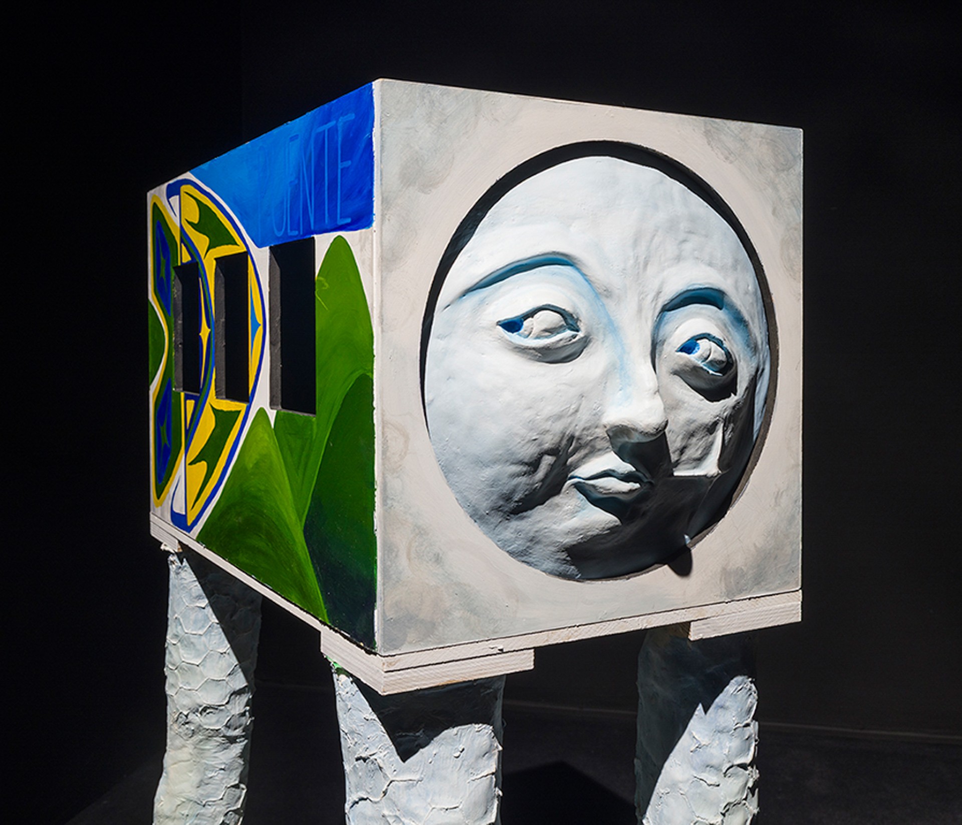 Une sculpture représentant un bus coloré, doté de longues pattes blanches et d'un visage énigmatique. La sculpture se détache sur la pièce noire dans laquelle elle se trouve.