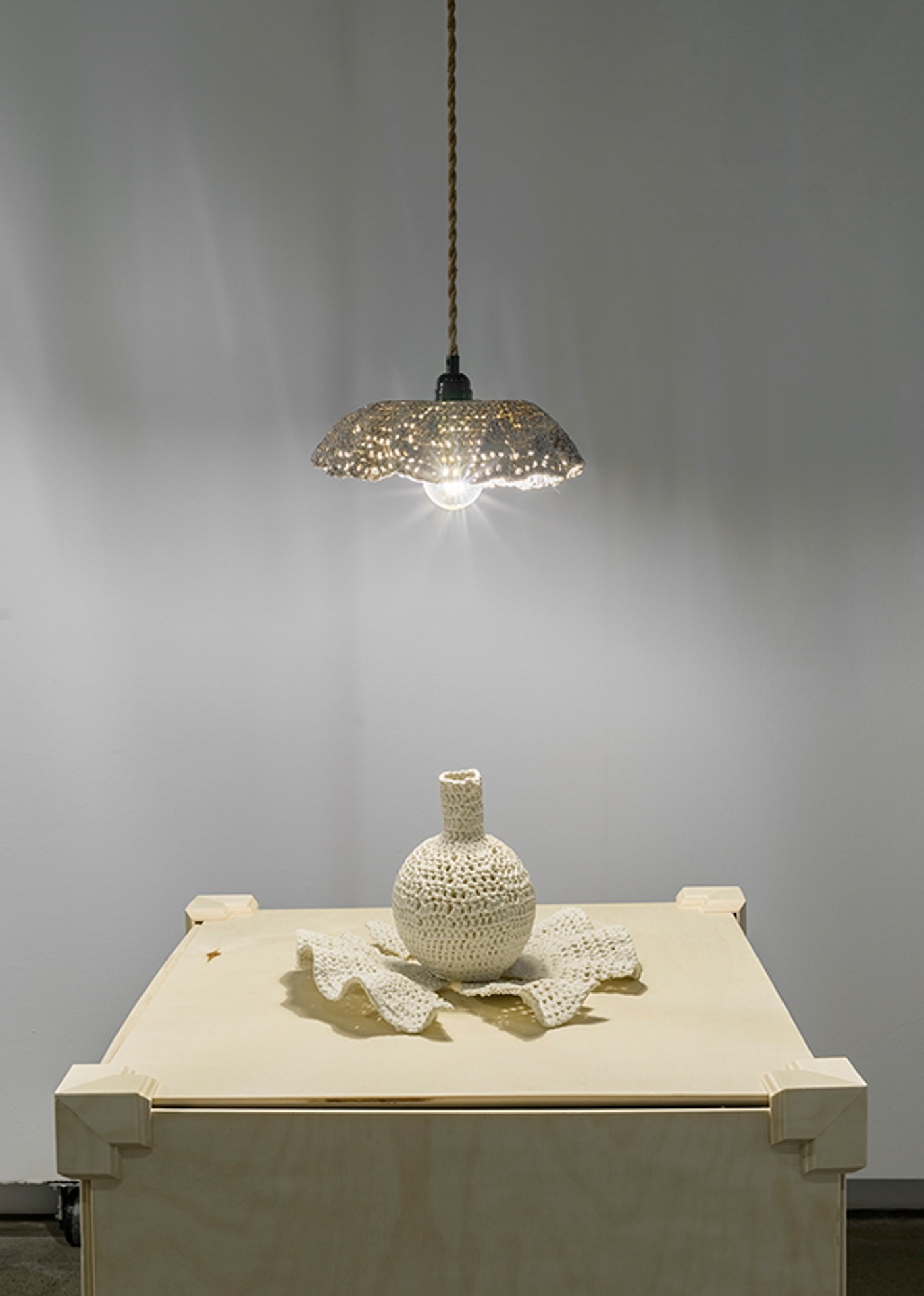 un vase en porcelaine exposé, qui ressemble à un ouvrage au crochet, posé sur un socle réglable. Il est éclairé par une suspension également en porcelaine, qui semble elle aussi avoir été réalisée au crochet, et dont la lumière transparaît à travers les mailles.