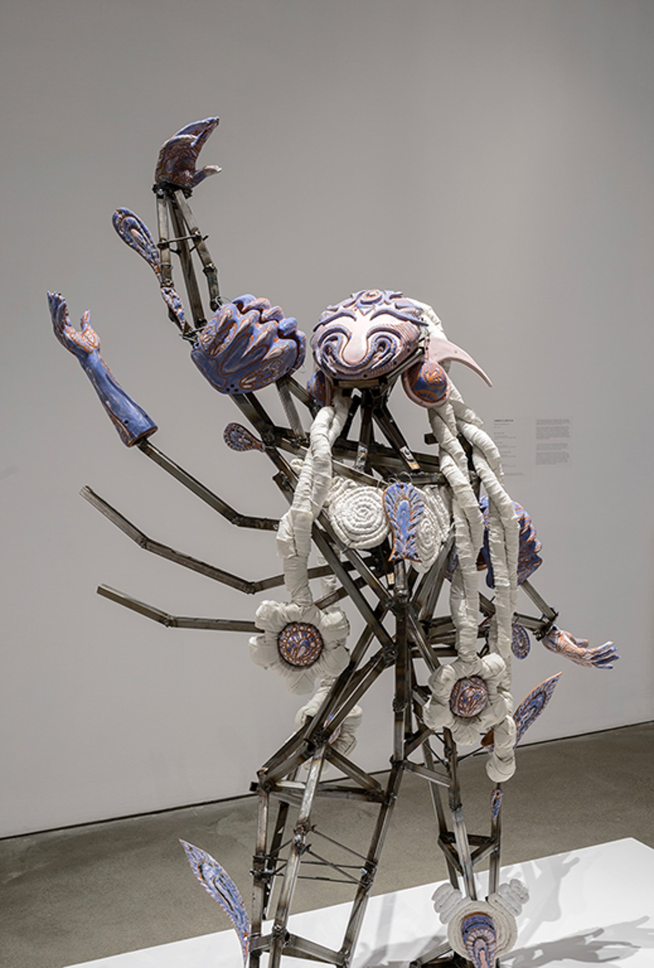 Sculpture représentant une figure robotique composée de pièces métalliques et textiles, l'un de ses quatre bras levé vers le ciel. 