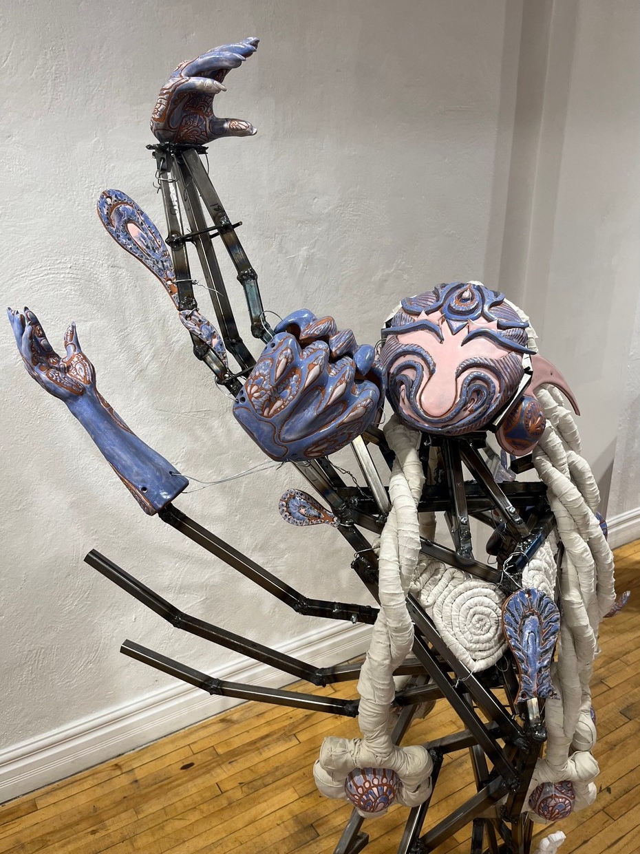 Sculpture représentant une figure robotique composée de pièces métalliques, dont le visage est réalisé en céramique violette et rose avec des détails ornés et des tresses textiles.