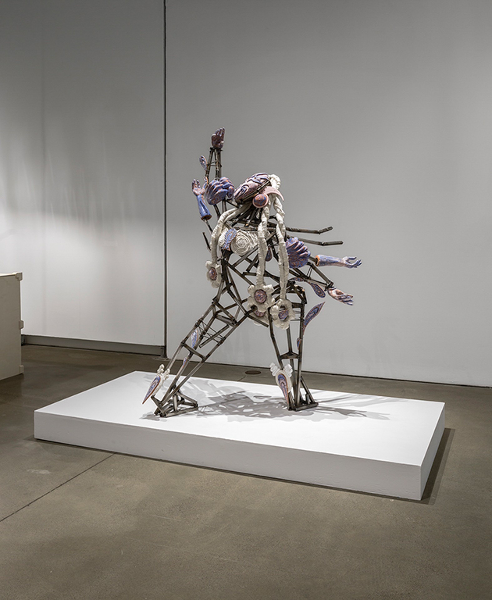 Sculpture représentant une figure robotique composée de pièces métalliques, dont le visage est réalisé en céramique violette et rose avec des détails ornés et des tresses textiles.