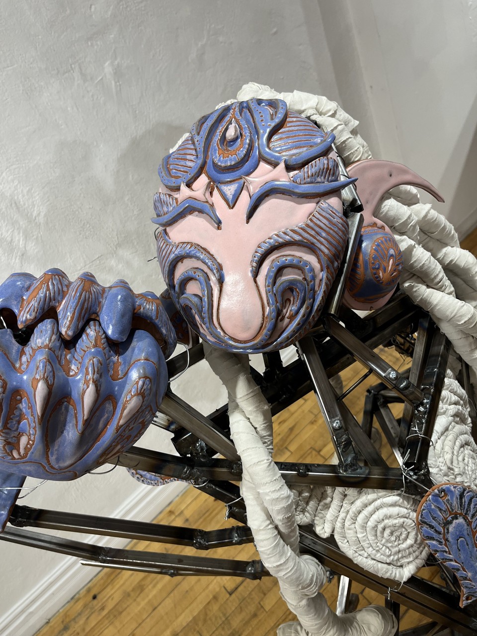 Sculpture représentant une figure robotique composée de pièces métalliques, son visage est fait de céramique violette et rose avec des détails ornés et des tresses en tissu blanc.