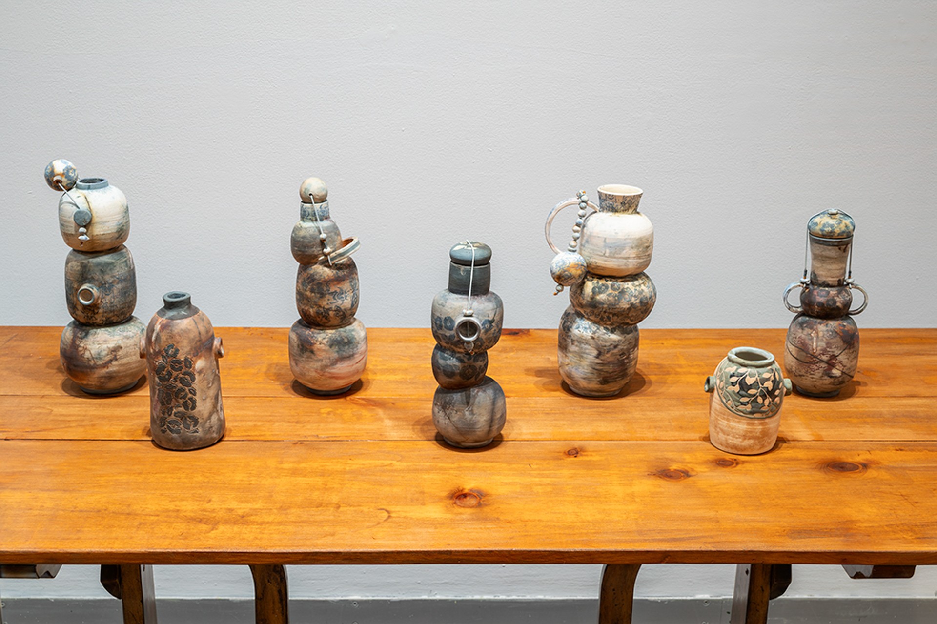 Une table en bois sur laquelle sont posés plusieurs vases en céramique. Ces vases sont tous composés de multiples formes arrondies ; certains sont dotés de couvercles suspendus, d'autres présentent des motifs, et d'autres encore sont munis d'anses.