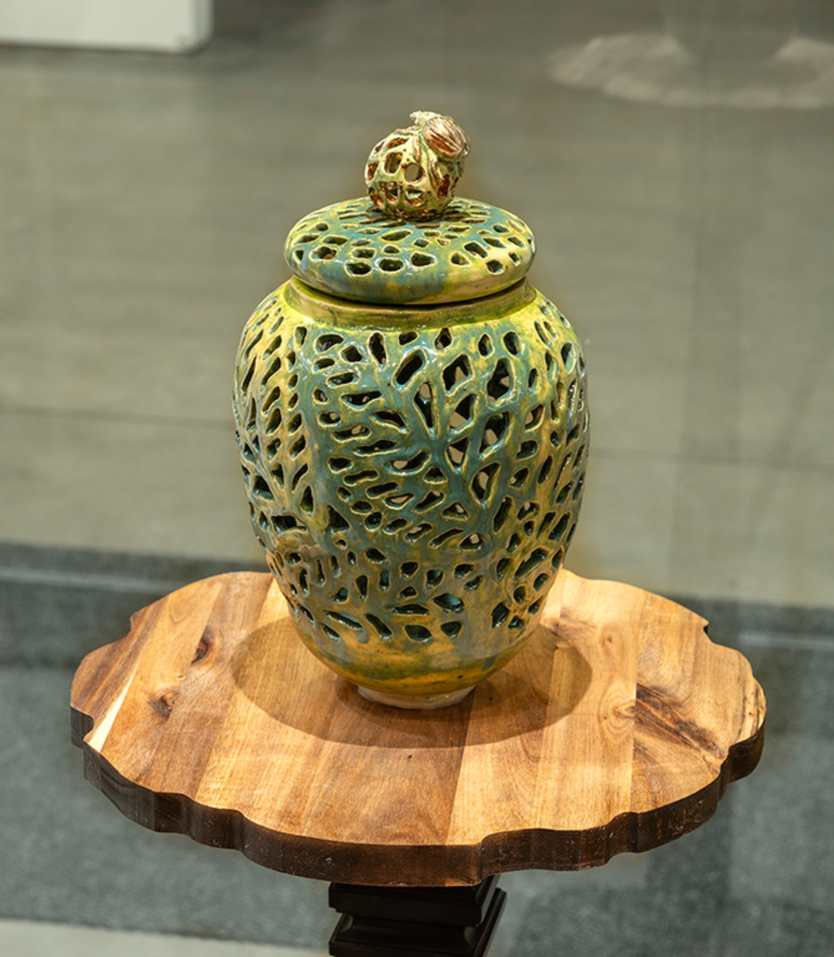 Un vase en céramique vert et jaune, orné de motifs en forme de feuilles. Le vase est posé sur une petite table d'exposition en bois.