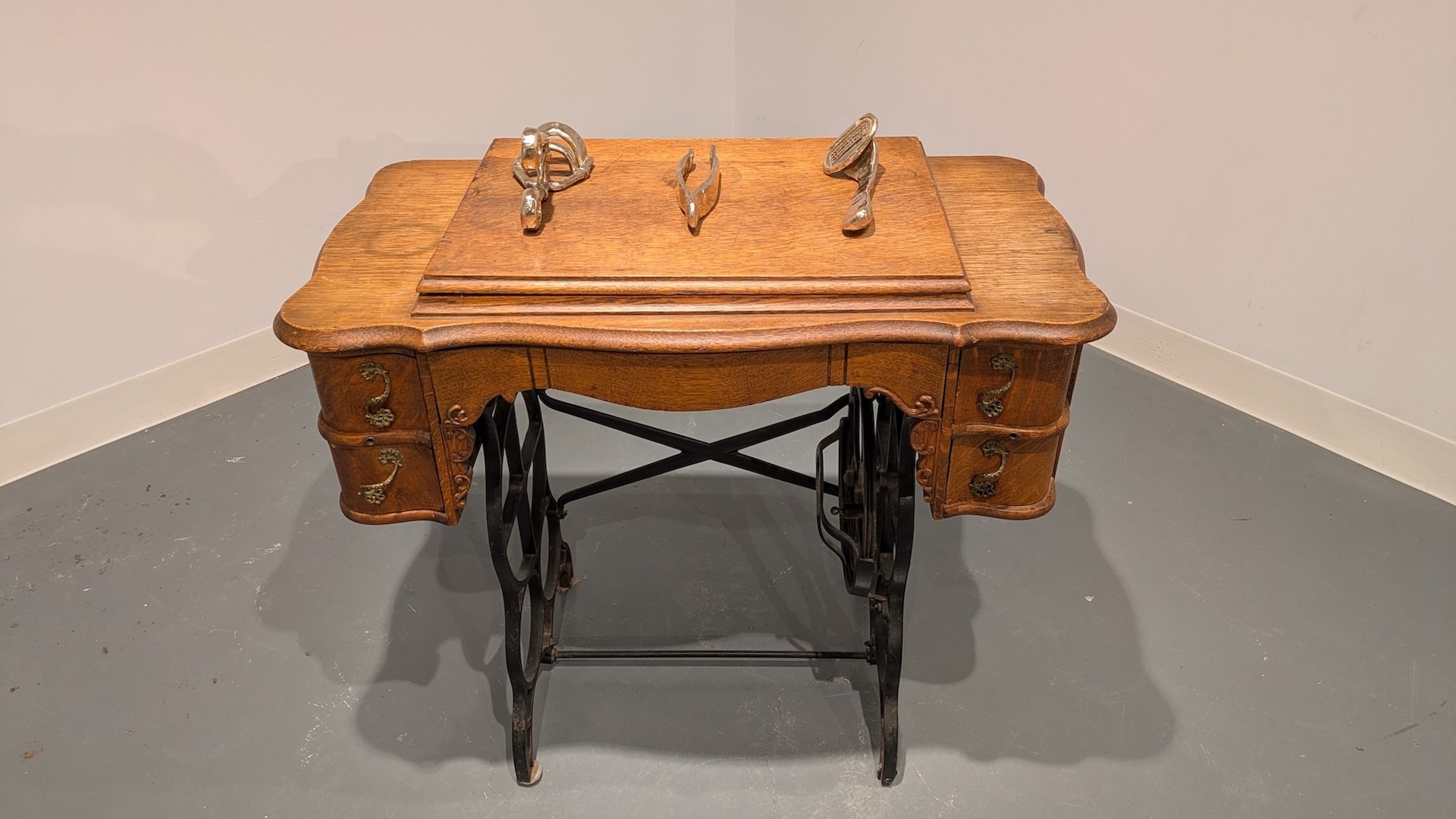 Table à couture décorée et avec trois objets surdimensionnés en bronze posés sur sa surface : un recourbe-cils, une pince à épiler et un rasoir jetable.