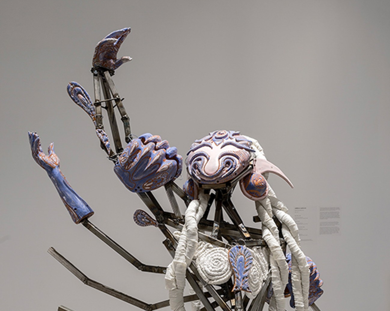 Sculpture représentant une figure robotique composée de pièces métalliques et textiles, l'un de ses quatre bras levé vers le ciel. 