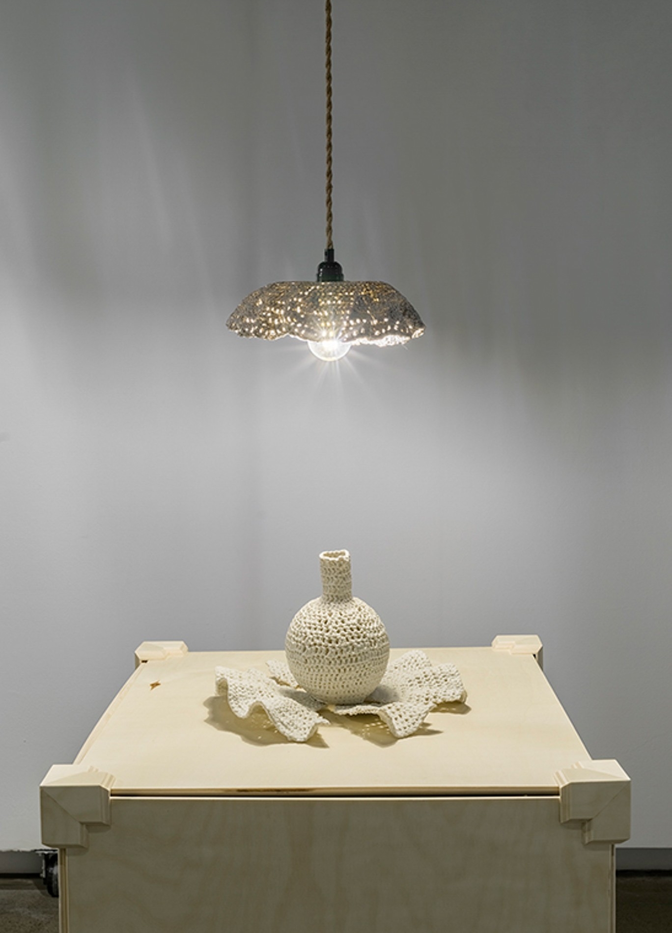 Une suspension est accrochée au-dessus d'un socle en bois sur lequel sont posés un vase rond et une assiette cassée ; l'abat-jour, le vase et l'assiette sont en porcelaine et ressemblent à du crochet