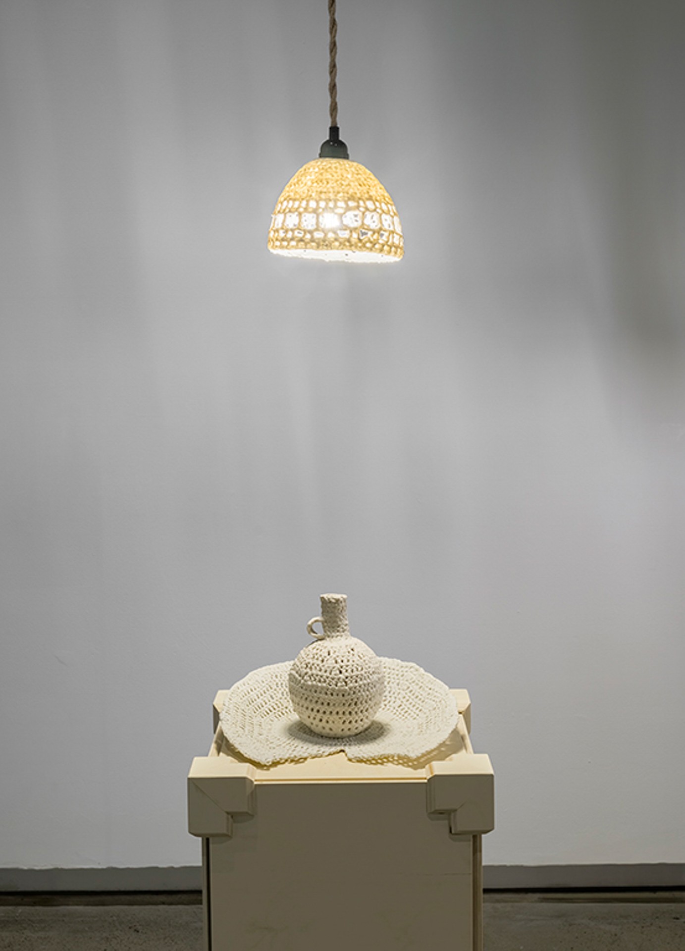 Une suspension est accrochée au-dessus d'un socle en bois sur lequel sont posés un vase rond et une assiette cassée ; l'abat-jour, le vase et l'assiette sont en porcelaine et ressemblent à du crochet