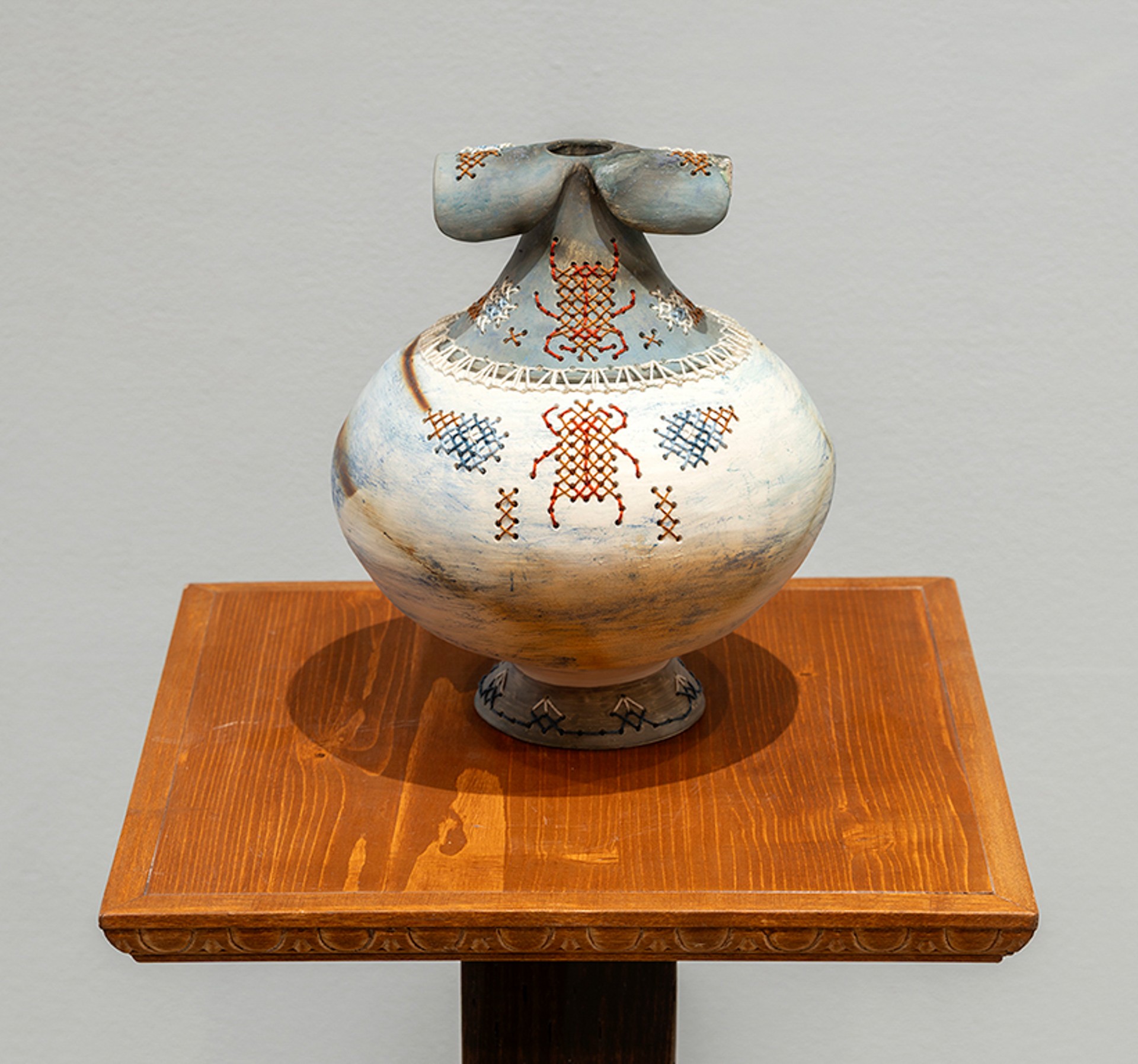 Sur une base en bois: un vase en céramique bleu et blanc, décoré de scarabées rouges brodés et de fleurs bleues, blanches et rouges.