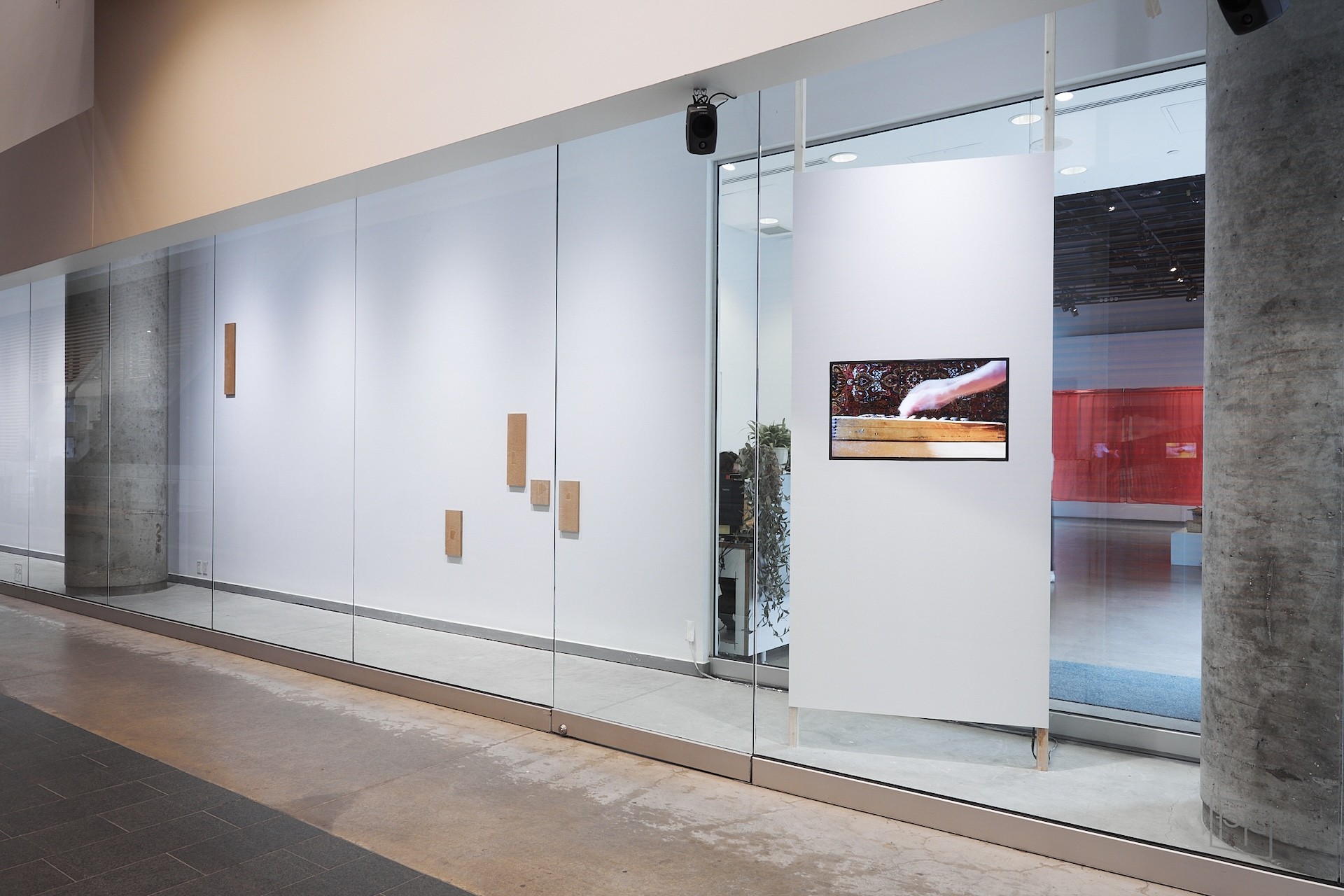 Vue d’installation de deux œuvres dans une vitrine de couloir. L’une présente une vidéo figée montrant une main manipulant un boulier. La vidéo est projetée sur un long mur vertical, orienté différemment du couloir et de la vitrine. À gauche se trouve le mur blanc habituel, ponctué de sculptures rectangulaires en bois. 