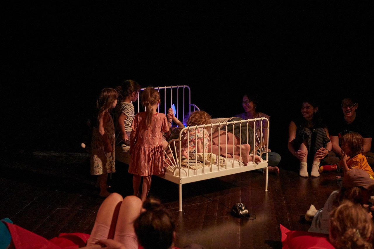Vue d’une galerie aux murs noirs, avec plusieurs jeunes enfants assis sur un petit lit ou y grimpant, doté d’une tête et d’un pied de lit en métal. La scène est éclairée d’une lumière chaude, contrastant avec l’obscurité dans laquelle se trouve le public.