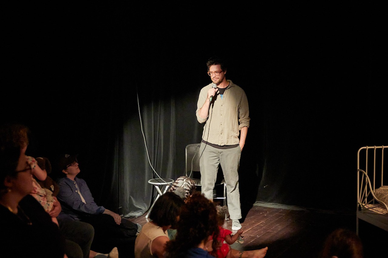 Vue d’une galerie aux murs noirs, avec Dillon de Give debout au centre de l’espace de performance, tenant un microphone et éclairé par un projecteur. Il semble avoir interrompu son discours et regarde le public en souriant.