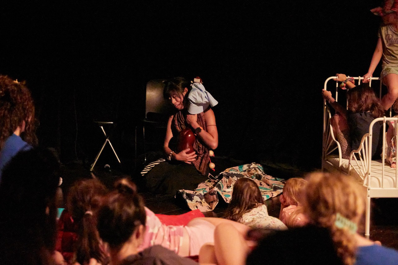 Vue d’une performance autour d’une routine du coucher, avec la performeuse Ansona Ching assise au sol, entourée d’une couverture, tenant un instrument et un chapeau qui représente son fils. Elle et les enfants qui l’entourent dans la pièce sombre sont éclairés.