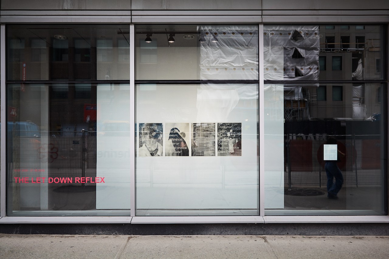 Vue extérieure de la vitrine de la galerie FOFA depuis la rue Sainte-Catherine. Le titre de l’exposition, « The Let Down Reflex », est inscrit en rose dans la partie inférieure gauche de la vitrine. Le reflet d’une construction urbaine est visible dans la vitre.