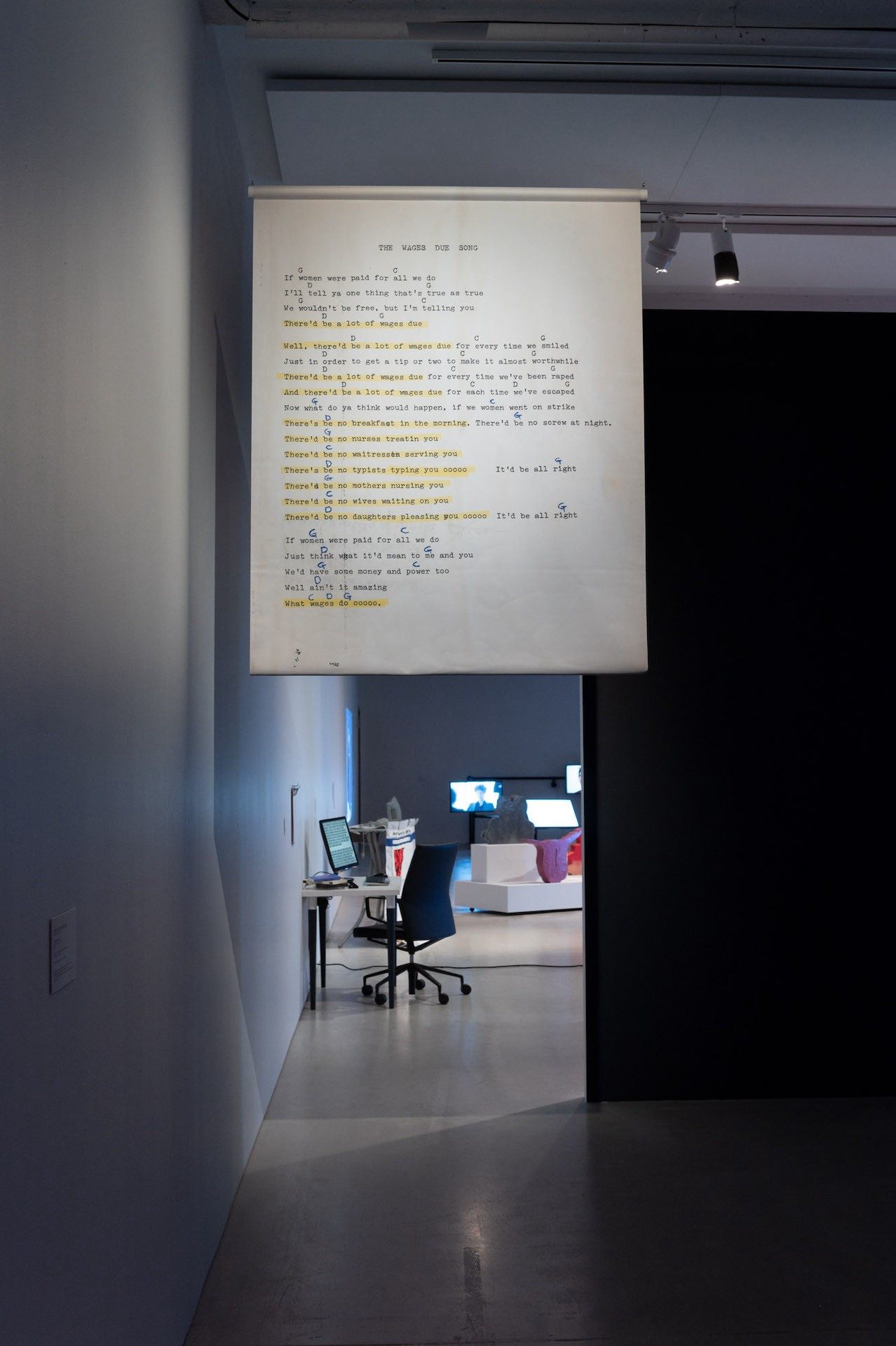 An installation view of a banner of the lyrics of Bo Watson's "Wages due song" with parts that are highlighted in yellow. Behind the installation, more artworks are visible in the gallery, including an installation of a working desk and several screens.