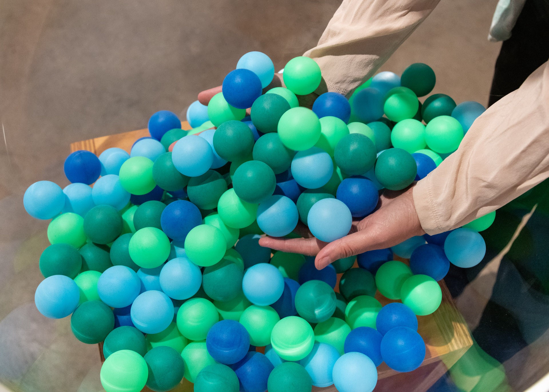 On voit les deux bras d'une personne s'étendre vers la droite du cadre pour retenir de nombreuses balles de ping-pong vertes et bleues provenant d'un bol posé sur un socle. Les balles de ping-pong commencent à déborder sur les côtés.