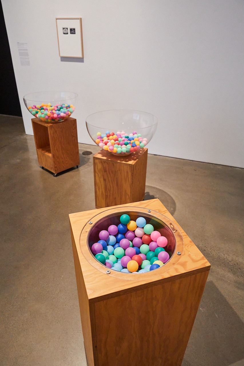 Une installation dans une galerie d'art composée de trois socles de conception différente sur lesquels sont disposées des balles de ping-pong multicolores. Les œuvres sont exposées dans une galerie aux murs blancs.