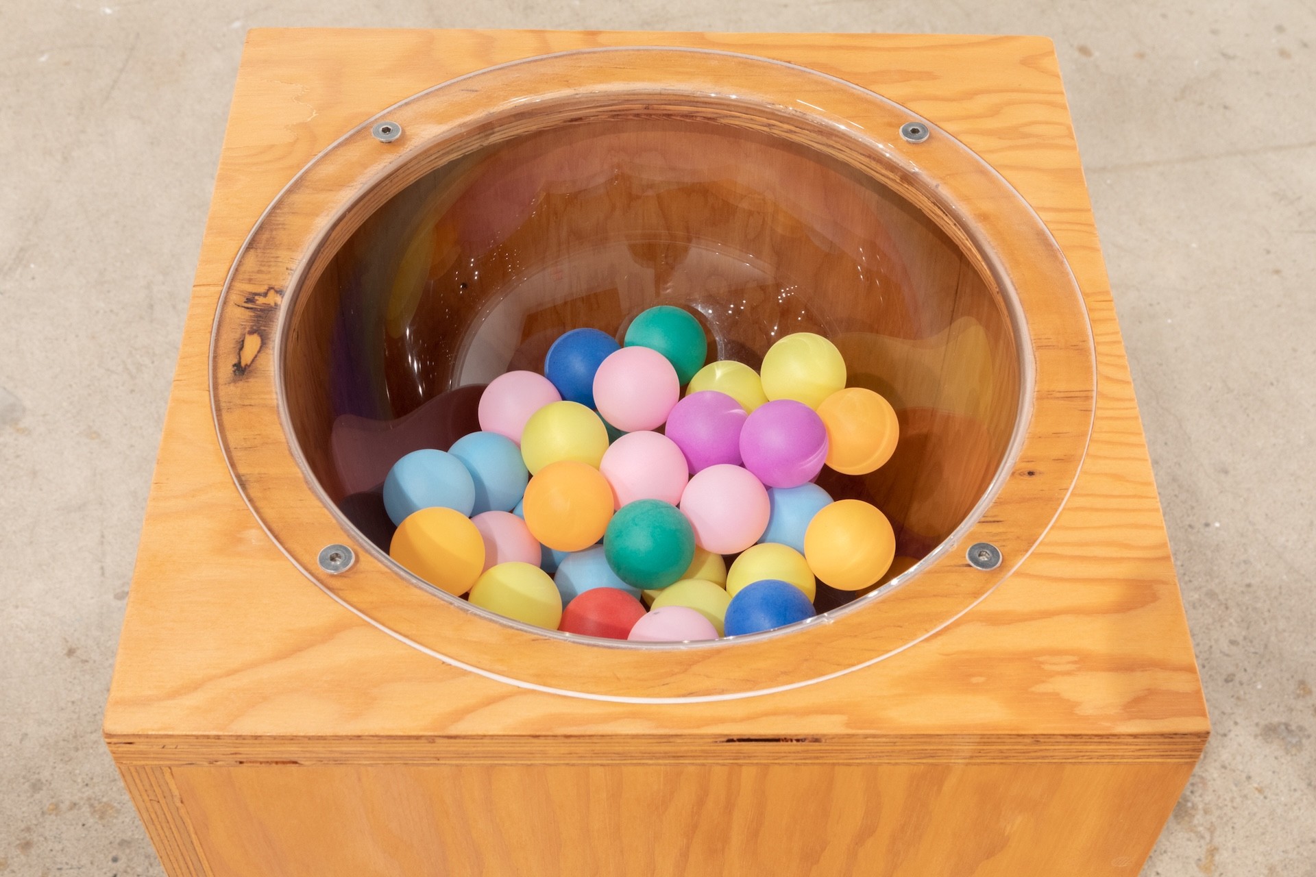 Gros plan sur un socle dans lequel est encastré, au centre, un bol transparent. Le bol est rempli de balles de ping-pong multicolores.