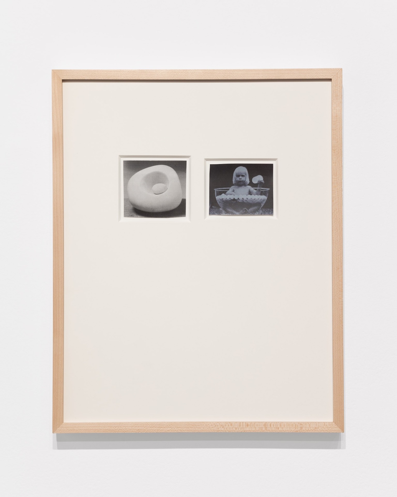 Un mur blanc sur lequel est accrochée une œuvre d'art. Celle-ci est encadrée dans du bois clair et comporte deux images plus petites à l'intérieur. L'une représente une forme pâle, arrondie et sculpturale ; l'autre, un bébé assis dans un bol d'eau rempli de balles de ping-pong.