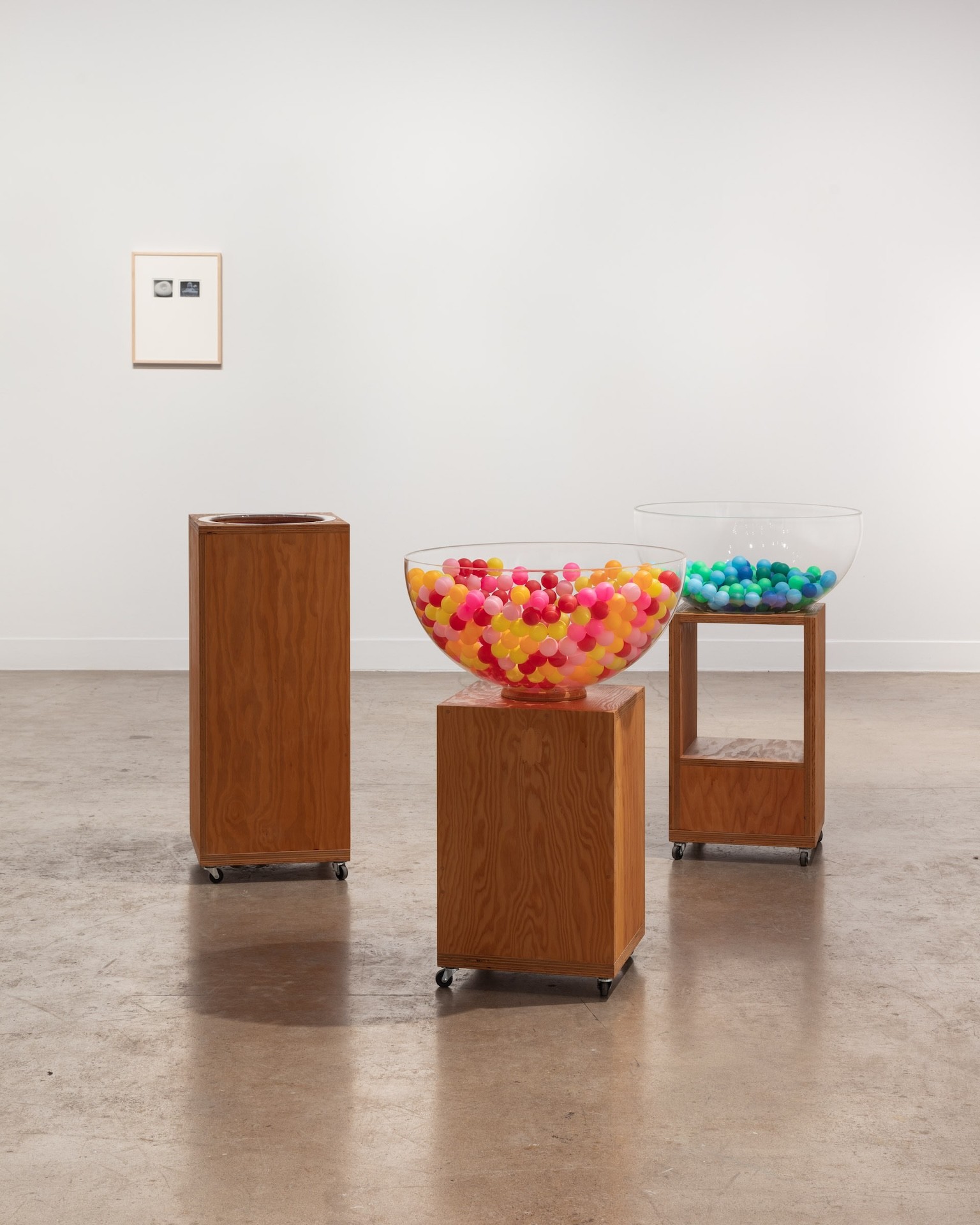 Une installation dans une galerie d'art composée de trois socles de conception différente sur lesquels sont disposées des balles de ping-pong multicolores. Les œuvres sont exposées dans une galerie aux murs blancs.