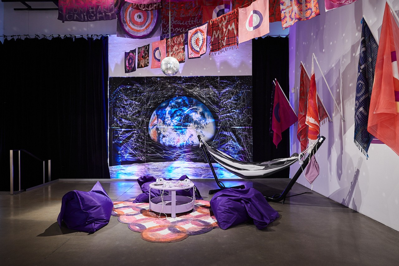 Une installation en galerie d’art comprenant une affiche, plusieurs drapeaux fixés aux murs et suspendus à des supports au plafond. Un hamac est placé au centre de la pièce, le sol est recouvert d’un tapis violet, et à l’arrière, une immense affiche murale représente une planète néon.