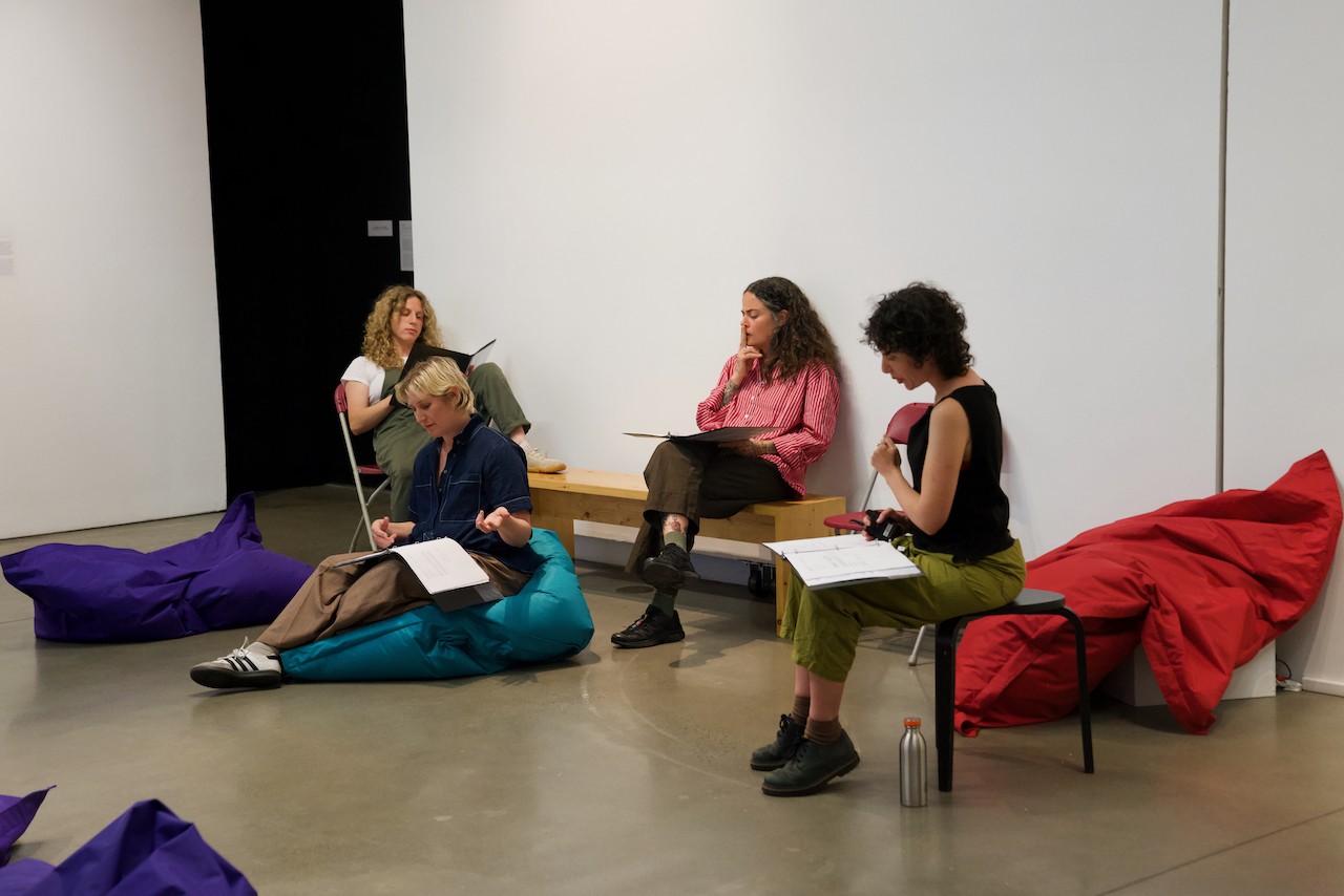 Vue de trois performeuses enceintes assises sur de grands poufs dans un espace de galerie. Elles ont chacune le script ouvert devant elles. Rachel est assise sur un tabouret et les regarde. L’une des performeuses est figée en pleine prise de parole, la main levée pour appuyer son propos.