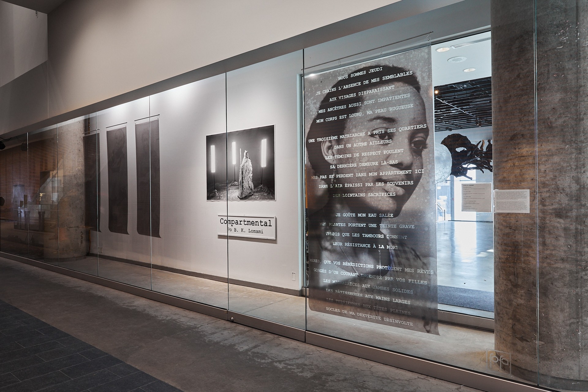 Vue d'une installation dans une vitrine composée de six œuvres : de gauche à droite, trois photos verticales suspendues au plafond sur un film opaque, représentant chacune trois femmes ; une photo de paysage montrant un personnage vêtu de papier d'aluminium, entouré de trois puissants projecteurs ; un panneau portant l'inscription « Compartmental. Po B. K. Lomami », et une grande photo sur film translucide suspendue au plafond représentant une femme avec un poème juxtaposé ; à travers la pièce translucide, on aperçoit l'espace ouvert de la galerie