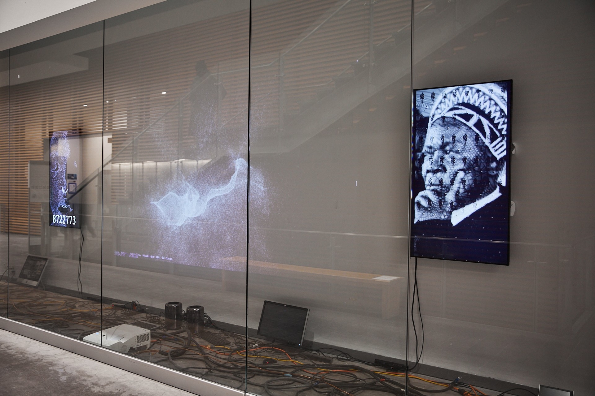 Vue d'une installation présentée dans une vitrine, composée de trois œuvres d'art avec des câbles et divers équipements électroniques disposés au sol, de gauche à droite : un écran vertical affichant un visage pixélisé avec, en dessous, un compte à rebours arrêté à « 8722773 » ; une grande projection panoramique représentant un nuage amorphe de points sur fond noir ; et un écran vertical présentant le portrait en noir et blanc d'un homme noir pensif, portant un couvre-chef, avec de petites figures juxtaposées