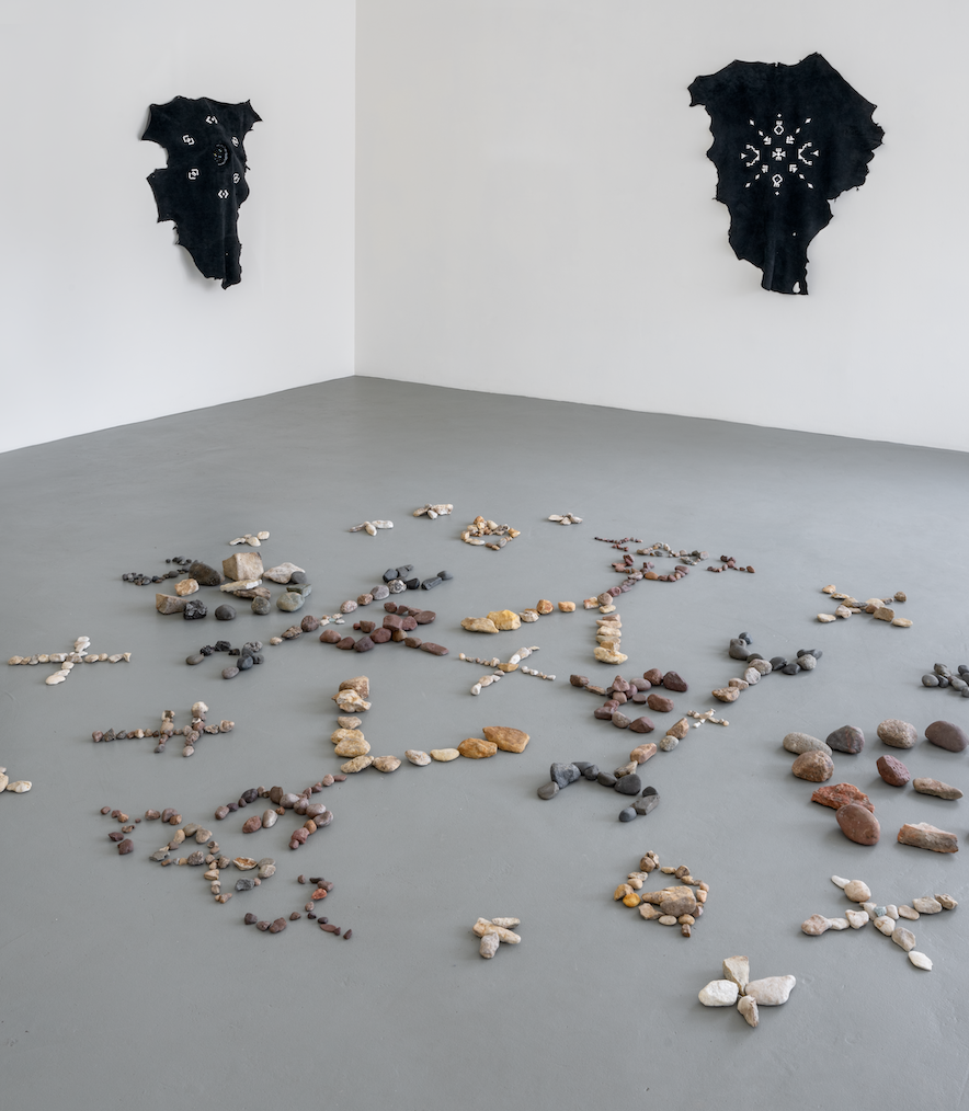 Sur le sol gris d’une galerie à espace blanc, une installation de pierres forme des motifs géométriques. Sur les murs sont suspendues deux peaux brodées.