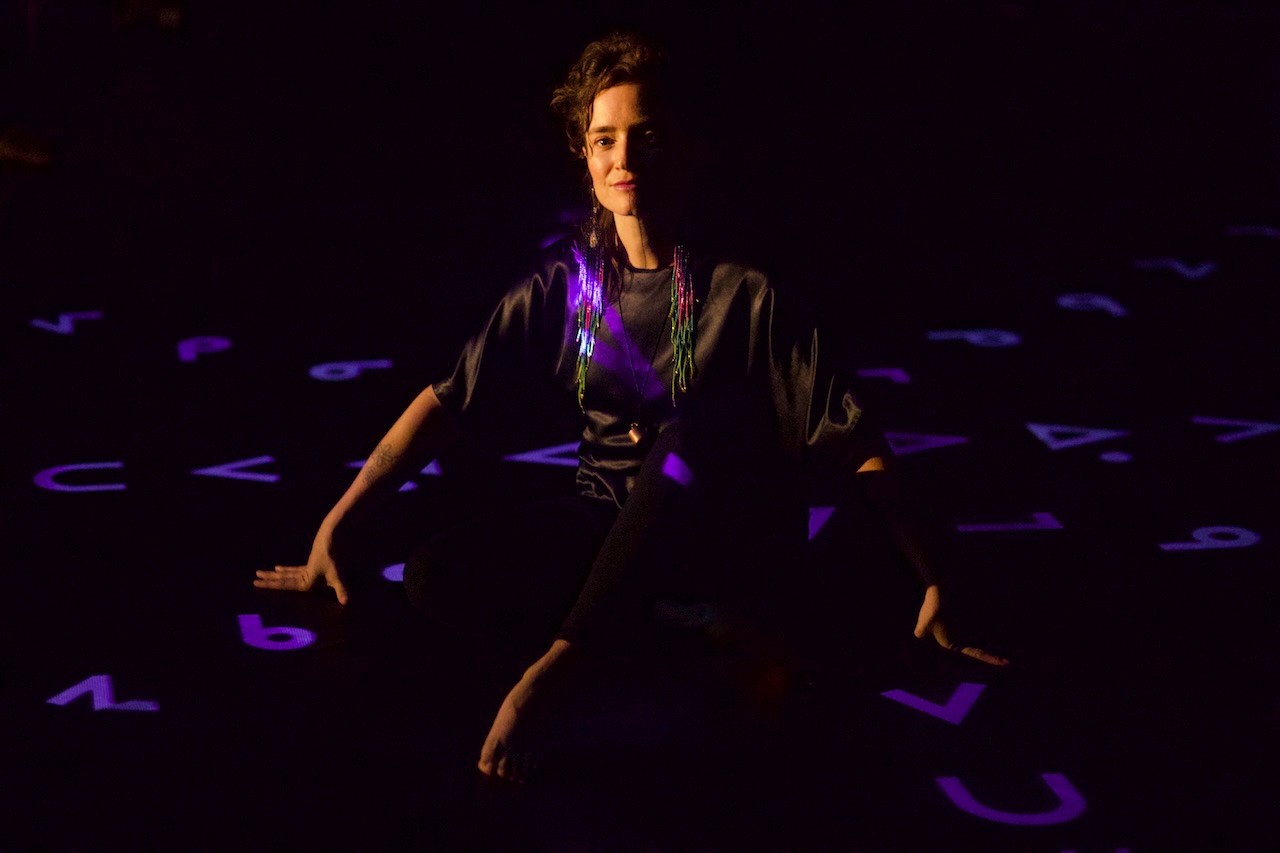 Moe Clark assise sur le sol d’une salle noire, entourée de projections violettes de caractères cris sur les murs et le sol. Ses mains sont posées au sol pour soutenir son corps.