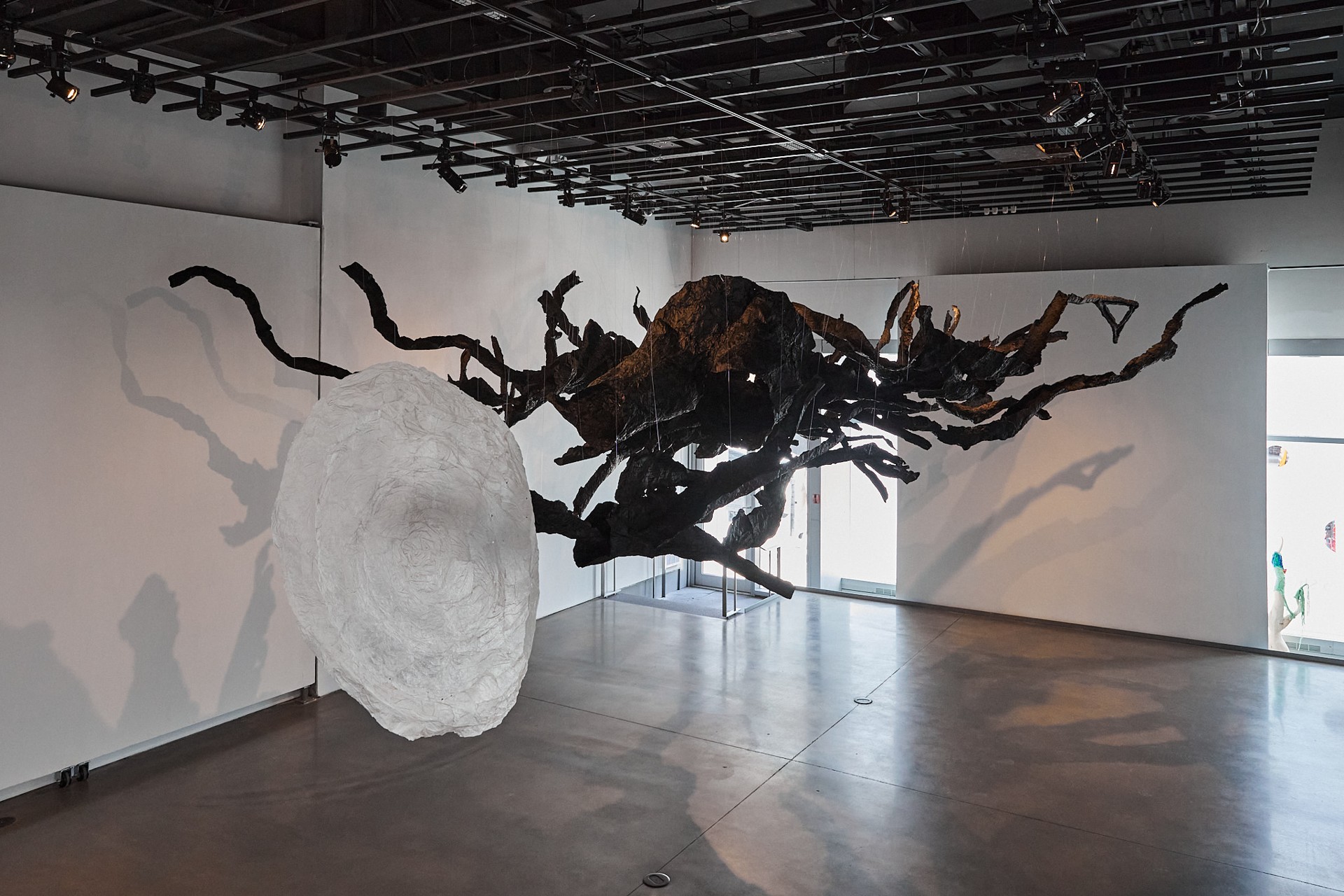 Vue de l'exposition montrant une grande galerie ouverte où est suspendue une sculpture noire évoquant un ensemble informe de branches d'arbres, avec, devant elle, un cercle de papier blanc suspendu à la même hauteur
