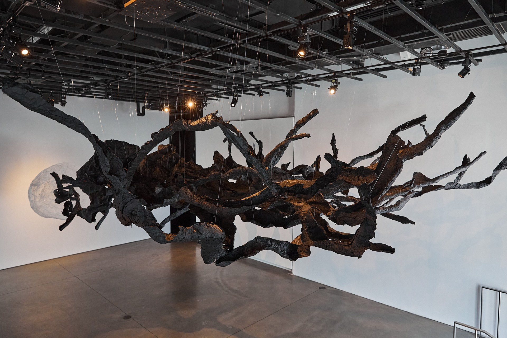 Vue de l'exposition montrant une grande galerie ouverte où est suspendue une sculpture noire évoquant un ensemble informe de branches d'arbres, avec, derrière, un cercle de papier blanc suspendu à la même hauteur.