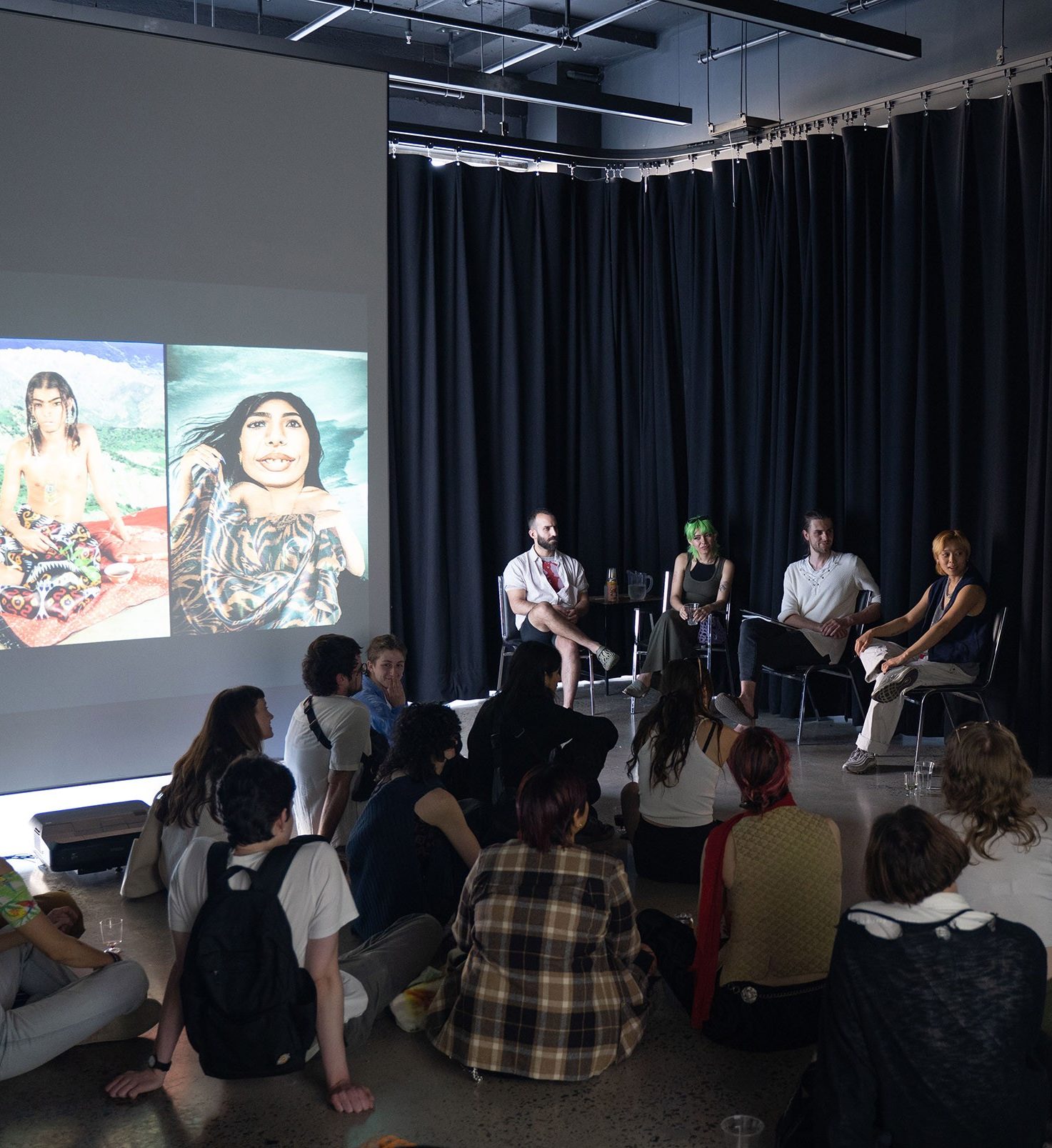 A panel of four people taking to an audience that is sitting on the floor.