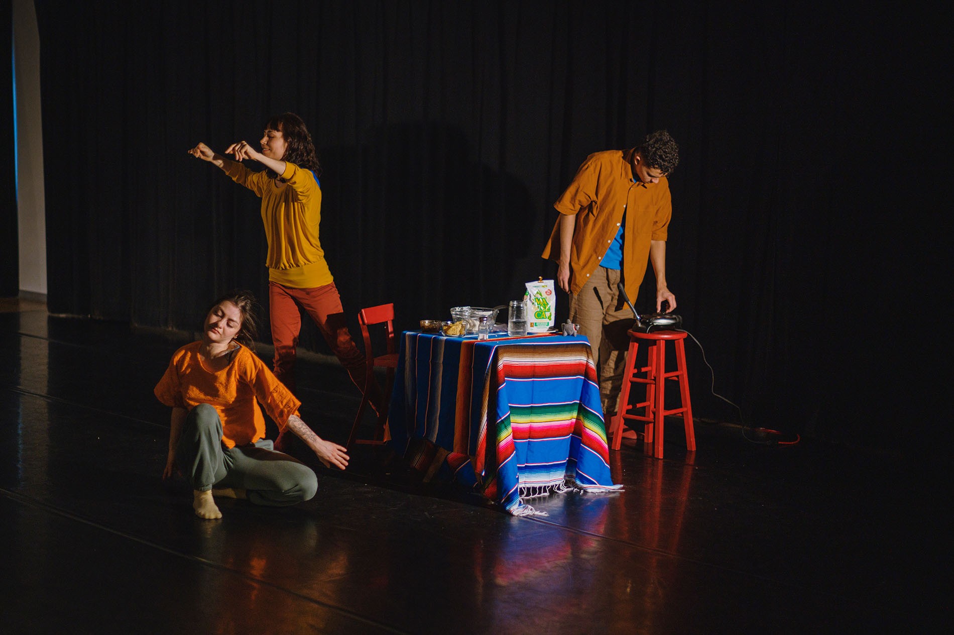 Three performers on stage with a table draped in a Mexican blanket 