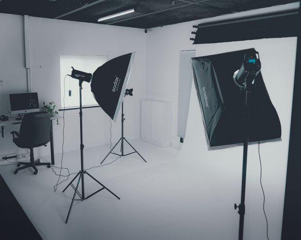 An image of a photography studio with a white backdrop, two soft box lights and a desk with a mac computer set up in the corner
