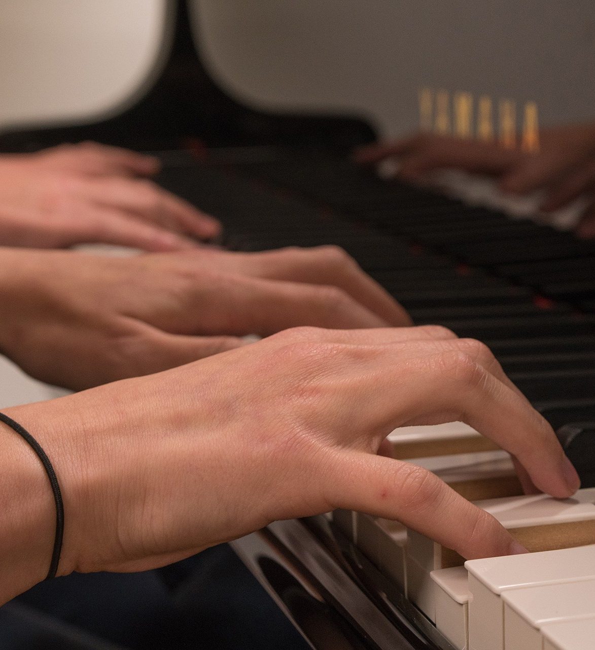 Close up of 4 hands on a piano keyboard; hands in the foreground are in focus.