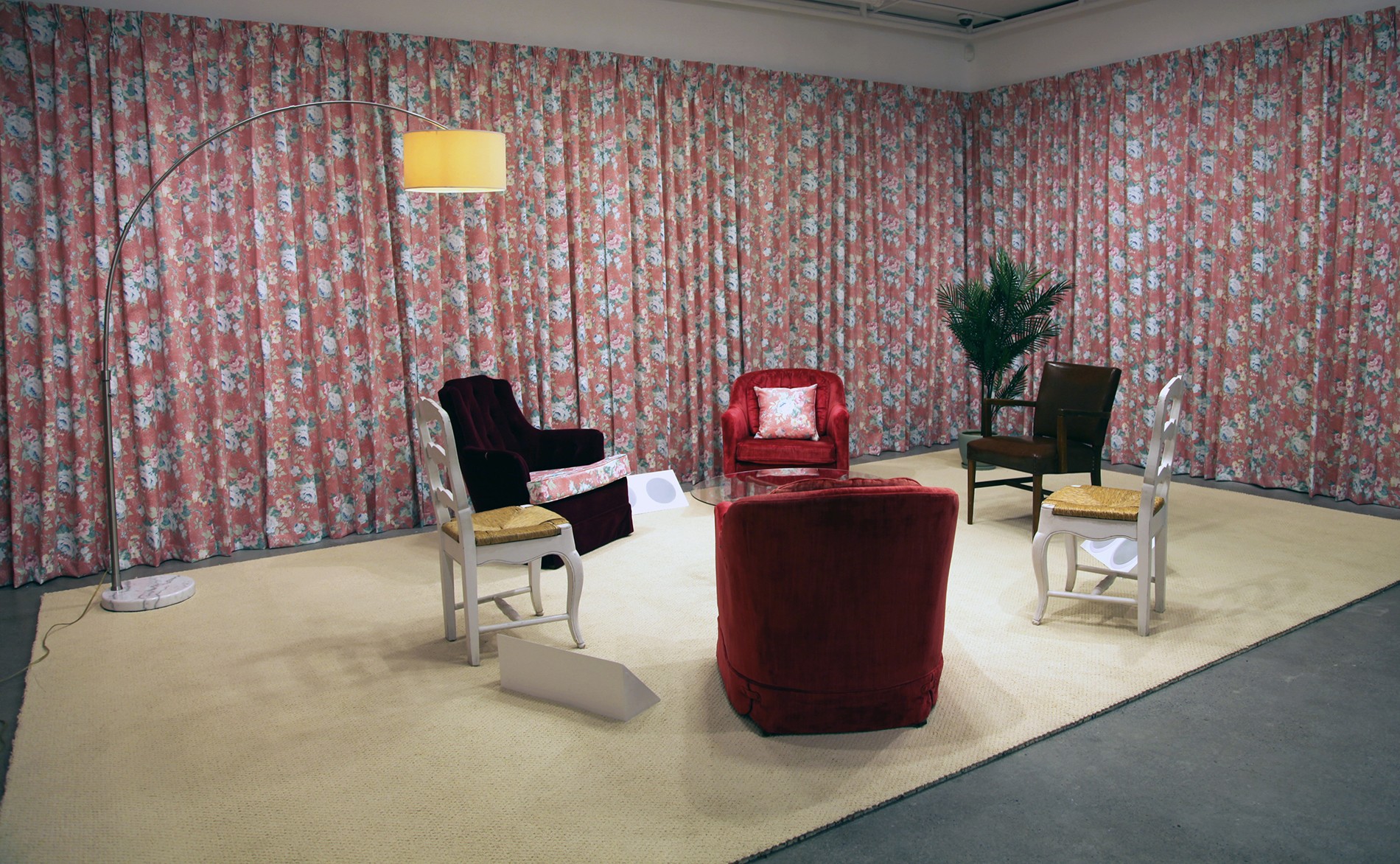 A room-like installation with floral-patterned curtains covering the walls. A light-colored rug covers the floor, on which several chairs are arranged in a circle. White speakers sit on the floor, tucked between the chairs. A tall, arched lamp stands to the left. A small plant is visible in the background. 