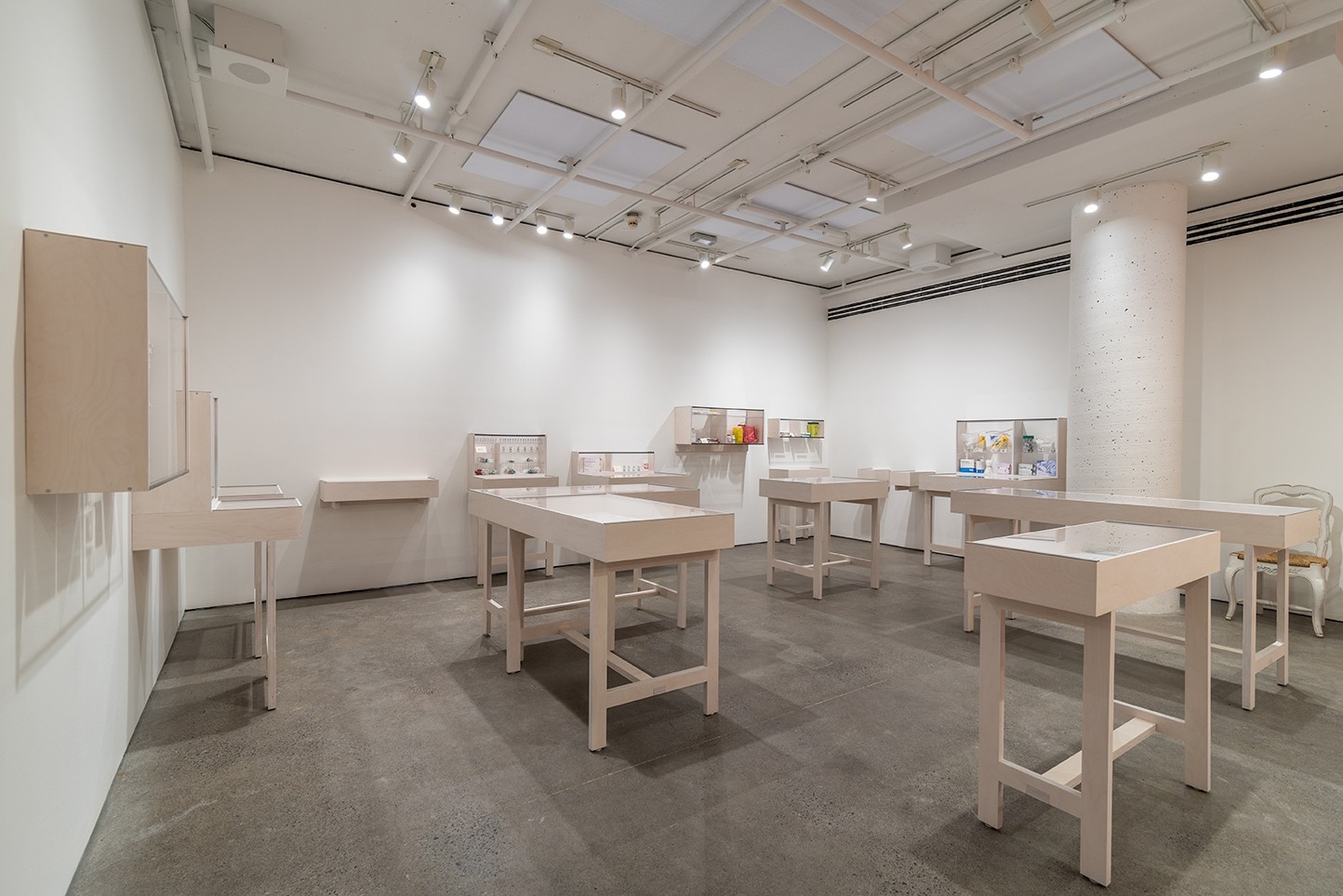 A brightly lit gallery space with white walls and a gray floor features numerous white display tables. The tables hold various small objects and artworks. Display cabinets are also mounted on the walls. Overhead, white pipes and lights are visible.