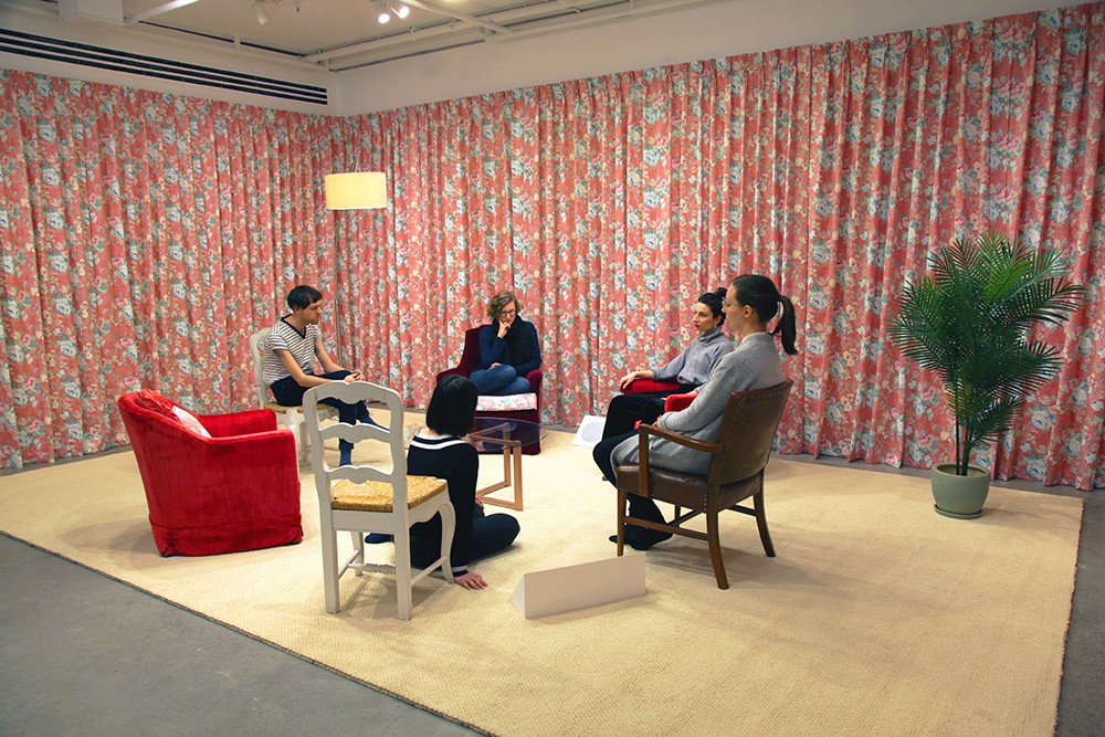 A room-like installation with floral-patterned curtains covering the walls. Five people are seated on chairs and the floor, arranged in a circle. A tall, arched lamp stands to the left. A small plant is visible on the right. It's not clear if the people are performers or visitors.