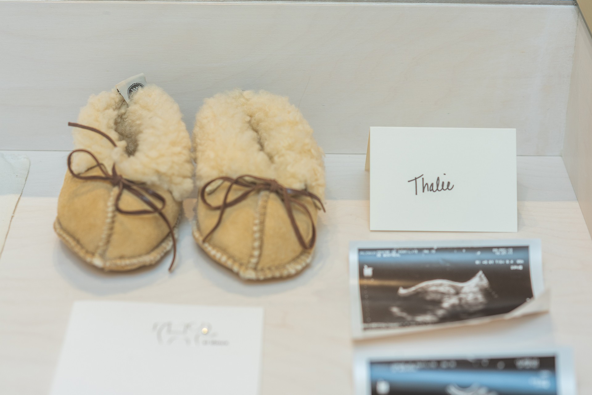 A close-up photograph shows a display case with a pair of soft, light-colored sheepskin slippers. A card with "Thalie" written on it is visible, along with ultrasound images.