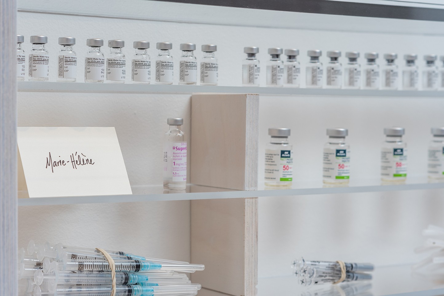 A close-up photograph shows a display case with rows of small vials and syringes on glass shelves. The vials appear to contain liquid. A card bearing the name "Marie-Hélène" rests on one of the shelves.