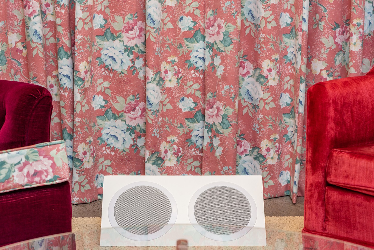 Close up of two white speakers, tucked between two velvet armchairs. A floral-print curtain frames the background of the installation.