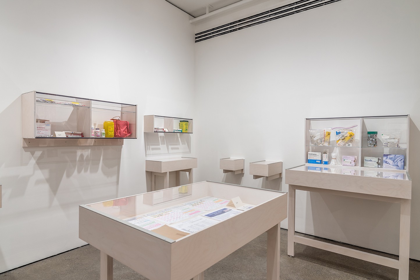 A brightly lit gallery space with white walls and a gray floor features white display cases and tables holding medical supplies or pharmaceuticals and their packaging. The objects are varied and colorful.