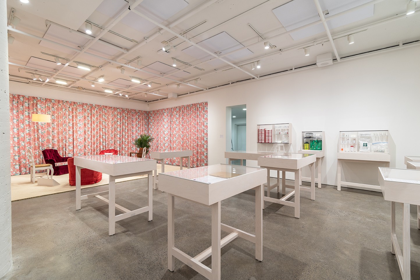 A brightly lit gallery space with white walls and a gray floor features white display tables holding small objects. Floral-patterned curtains line one wall, creating a distinct area with seating. Overhead pipes and lights are visible.
