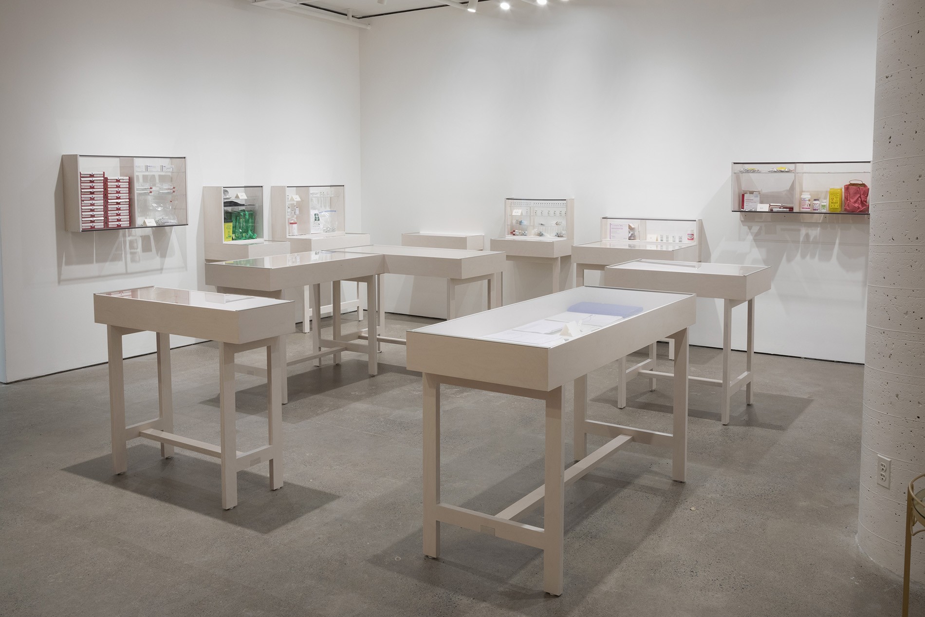 A brightly lit gallery space with white walls and a gray floor features white display tables holding small objects that are varied and colourful. The tables are arranged in a loose circle. 