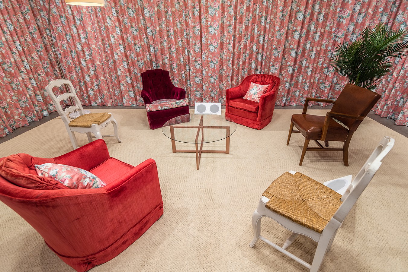 An overhead view of an installation representing a living room with 6 chairs and a circular glass coffee table. Floral-patterned curtains create walls for the space, and a two white speakers are positioned near the centre of the image, between two velvet armchairs.