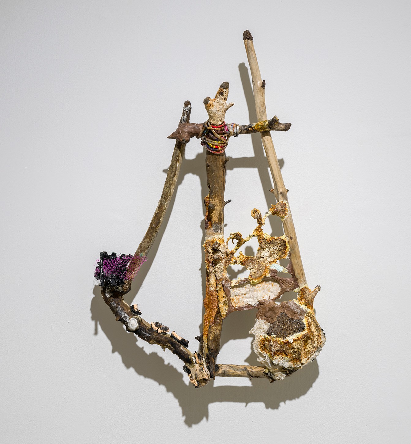 The trianglular sculpture composed sticks, yarn and colourful threads. The sculpture also includes a textured, golden-brown element and a purple, mesh-like material. Displayed on a white wall, the sculpture's shadow is cast behind it.