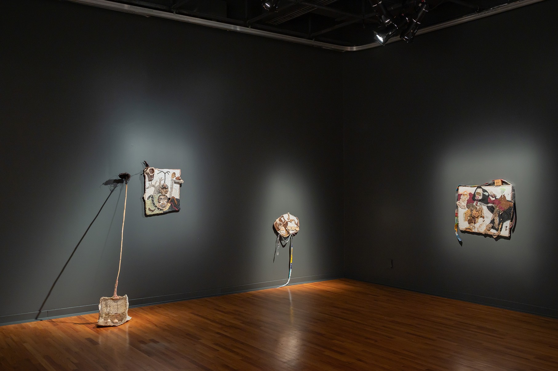A dimly lit gallery space with dark gray walls and a polished wooden floor features multiple artworks. Two square, textured artworks hang on the wall, one to the left and one to the right. A third, smaller artwork sits on the floor to the left, leaning against the wall. Spotlights on each piece create dramatic shadows